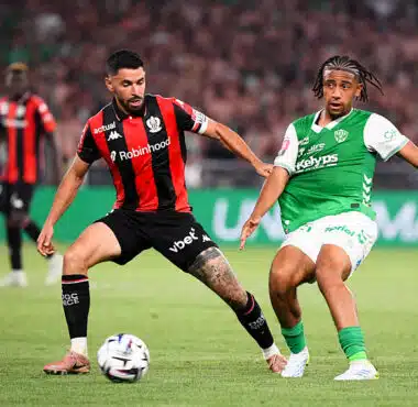 Two soccer players contesting the ball during a match; one wears red-and-black stripes, the other green-and-white kit, in a crowded stadium.