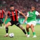 Two soccer players contesting the ball during a match; one wears red-and-black stripes, the other green-and-white kit, in a crowded stadium.