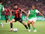 Two soccer players contesting the ball during a match; one wears red-and-black stripes, the other green-and-white kit, in a crowded stadium.