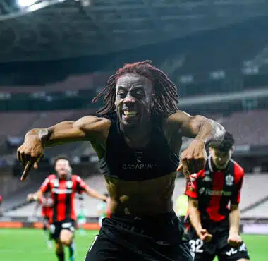 Soccer player with dreadlocks celebrates toward the camera on a stadium field, arms outstretched and smiling.