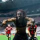 Soccer player with dreadlocks celebrates toward the camera on a stadium field, arms outstretched and smiling.