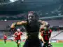 Soccer player with dreadlocks celebrates toward the camera on a stadium field, arms outstretched and smiling.