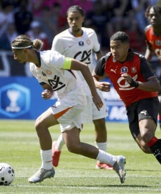 Soccer scene: a player in white with a green armband dribbles a ball while a defender in red and black chases closely from behind on a sunny grassy field.