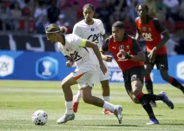 Soccer scene: a player in white with a green armband dribbles a ball while a defender in red and black chases closely from behind on a sunny grassy field.