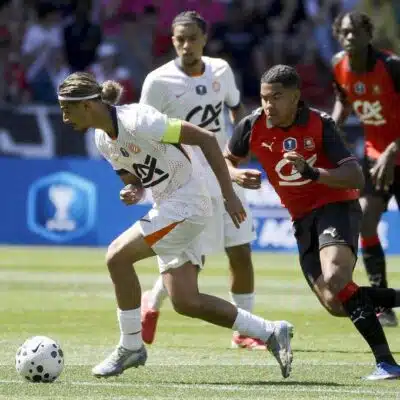 Soccer scene: a player in white with a green armband dribbles a ball while a defender in red and black chases closely from behind on a sunny grassy field.