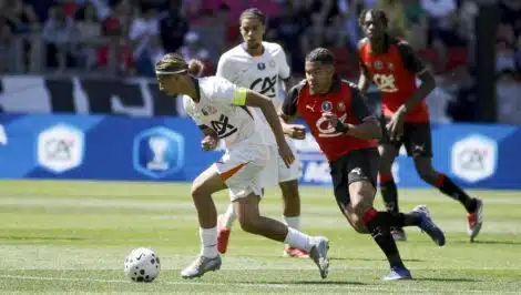 Soccer scene: a player in white with a green armband dribbles a ball while a defender in red and black chases closely from behind on a sunny grassy field.