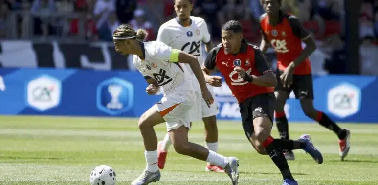 Soccer scene: a player in white with a green armband dribbles a ball while a defender in red and black chases closely from behind on a sunny grassy field.