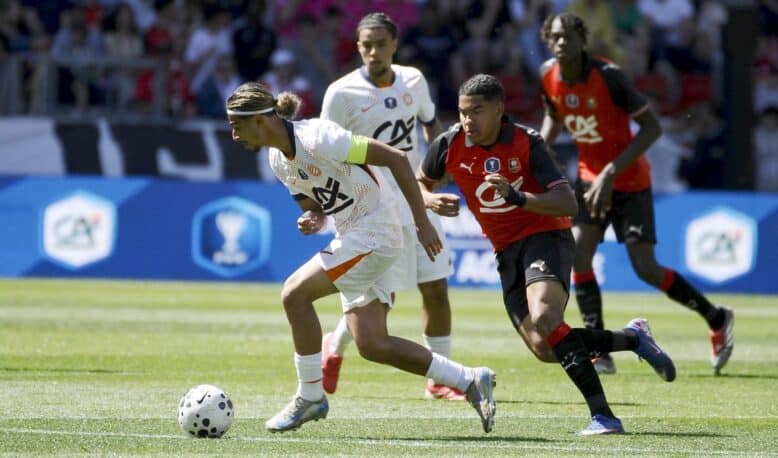 Soccer scene: a player in white with a green armband dribbles a ball while a defender in red and black chases closely from behind on a sunny grassy field.