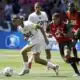 Soccer scene: a player in white with a green armband dribbles a ball while a defender in red and black chases closely from behind on a sunny grassy field.