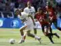 Soccer scene: a player in white with a green armband dribbles a ball while a defender in red and black chases closely from behind on a sunny grassy field.