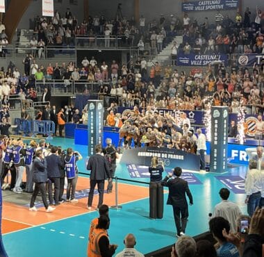 Large indoor sports arena filled with spectators watching a volleyball or similar match, with teams on a blue court and photographers nearby.