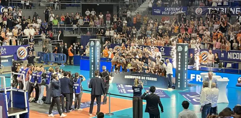 IMG_4226 | AllezPaillade.com Large indoor sports arena filled with spectators watching a volleyball or similar match, with teams on a blue court and photographers nearby.