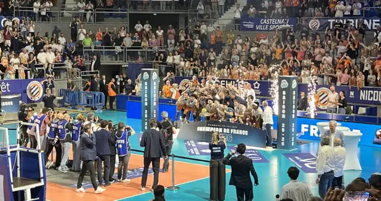 Large indoor sports arena filled with spectators watching a volleyball or similar match, with teams on a blue court and photographers nearby.
