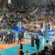 Large indoor sports arena filled with spectators watching a volleyball or similar match, with teams on a blue court and photographers nearby.