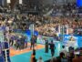 Large indoor sports arena filled with spectators watching a volleyball or similar match, with teams on a blue court and photographers nearby.