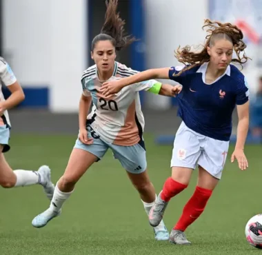 Three female soccer players sprint after the ball on a green field; one in a blue jersey with red socks leads the chase.