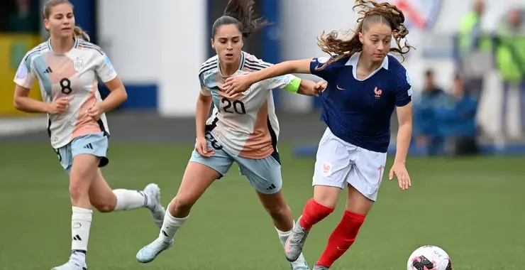 Three female soccer players sprint after the ball on a green field; one in a blue jersey with red socks leads the chase.