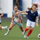 Three female soccer players sprint after the ball on a green field; one in a blue jersey with red socks leads the chase.