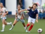Three female soccer players sprint after the ball on a green field; one in a blue jersey with red socks leads the chase.