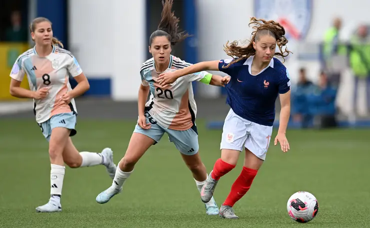 Three female soccer players sprint after the ball on a green field; one in a blue jersey with red socks leads the chase.