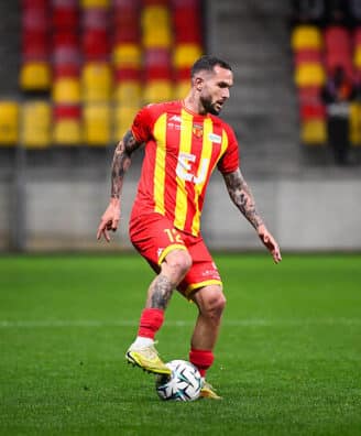 Soccer player in a red and yellow striped jersey dribbling a ball on a green field during a match.