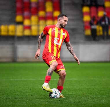 Soccer player in a red and yellow striped jersey dribbling a ball on a green field during a match.