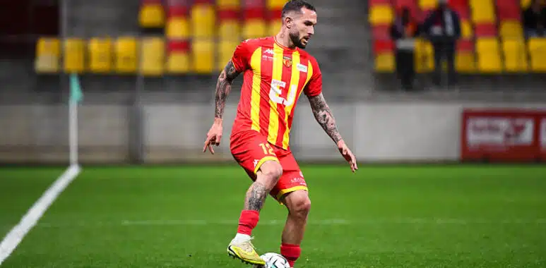 Soccer player in a red and yellow striped jersey dribbling a ball on a green field during a match.