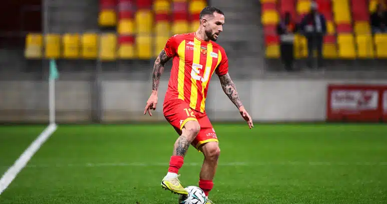 Soccer player in a red and yellow striped jersey dribbling a ball on a green field during a match.