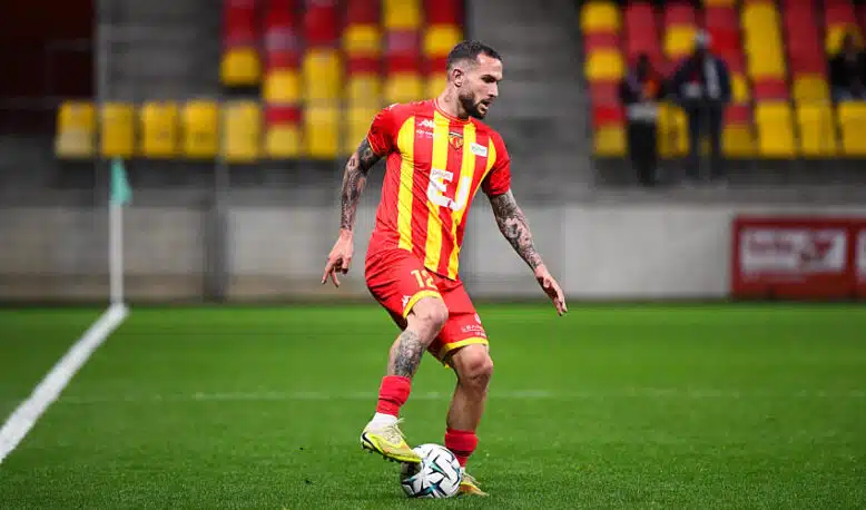 Soccer player in a red and yellow striped jersey dribbling a ball on a green field during a match.