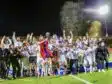 Screenshot | AllezPaillade.com Soccer team celebrates victory on a podium at night, lifting a trophy amid gold confetti.
