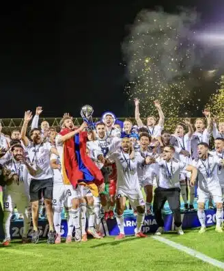 Soccer team celebrates victory on a podium at night, lifting a trophy amid gold confetti.
