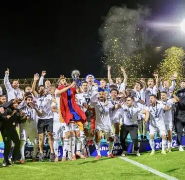 Soccer team celebrates victory on a podium at night, lifting a trophy amid gold confetti.
