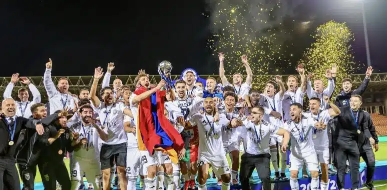 Soccer team celebrates victory on a podium at night, lifting a trophy amid gold confetti.