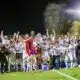 Soccer team celebrates victory on a podium at night, lifting a trophy amid gold confetti.