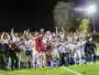 Soccer team celebrates victory on a podium at night, lifting a trophy amid gold confetti.