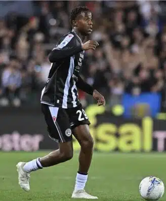 Screenshot | AllezPaillade.com Soccer player in a black and light-blue striped kit dribbles the ball on a green pitch with a cheering crowd in the background.
