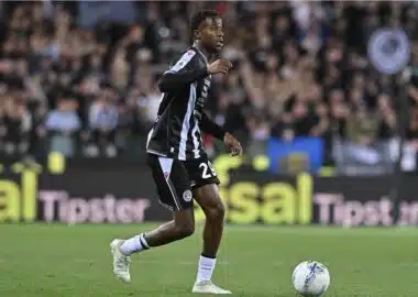 Soccer player in a black and light-blue striped kit dribbles the ball on a green pitch with a cheering crowd in the background.