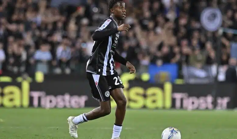Soccer player in a black and light-blue striped kit dribbles the ball on a green pitch with a cheering crowd in the background.