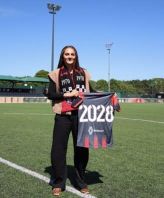 IMG_4706 | AllezPaillade.com Woman standing on a soccer field, holding a banner that reads 2028, wearing a black outfit with a red-and-black scarf.”