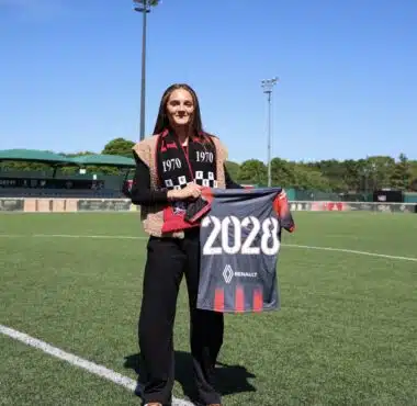 Woman standing on a soccer field, holding a banner that reads 2028, wearing a black outfit with a red-and-black scarf.&rdquo;