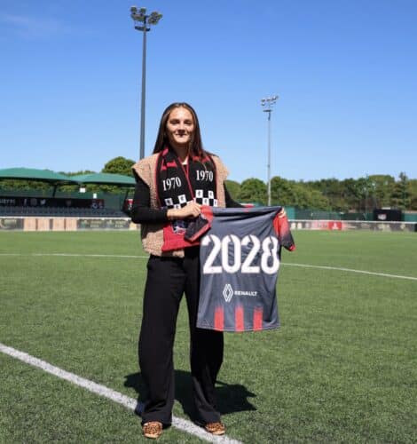 Woman standing on a soccer field, holding a banner that reads 2028, wearing a black outfit with a red-and-black scarf.&rdquo;