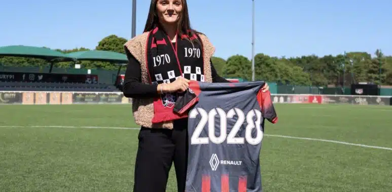 Woman standing on a soccer field, holding a banner that reads 2028, wearing a black outfit with a red-and-black scarf.&rdquo;