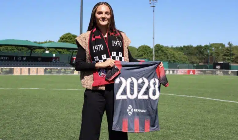 IMG_4706 | AllezPaillade.com Woman standing on a soccer field, holding a banner that reads 2028, wearing a black outfit with a red-and-black scarf.”