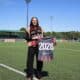 Woman standing on a soccer field, holding a banner that reads 2028, wearing a black outfit with a red-and-black scarf.&rdquo;