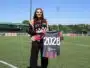 Woman standing on a soccer field, holding a banner that reads 2028, wearing a black outfit with a red-and-black scarf.&rdquo;