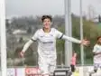 Young male soccer player in a white kit celebrates a goal with arms outstretched near the goalpost on a sunny day.