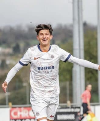 Young male soccer player in a white kit celebrates a goal with arms outstretched near the goalpost on a sunny day.