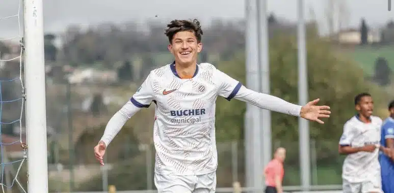 Young male soccer player in a white kit celebrates a goal with arms outstretched near the goalpost on a sunny day.