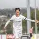 Young male soccer player in a white kit celebrates a goal with arms outstretched near the goalpost on a sunny day.