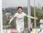 Young male soccer player in a white kit celebrates a goal with arms outstretched near the goalpost on a sunny day.